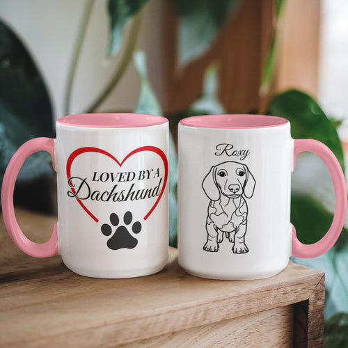 Two mugs with pink handles on a wooden surface, one featuring a heart and paw print design, the other with a dachshund illustration.