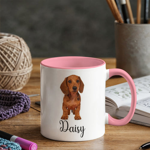 White mug with pink interior and handle featuring a dachshund illustration and 'Daisy' text, placed on a wooden surface with knitting materials.