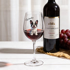 A clear wine glass with a Boston Terrier dog illustration and the name 'Winston' printed on it, filled with red wine, set on a white tabletop. A dark wine bottle and a bunch of red grapes appear blurred in the background.