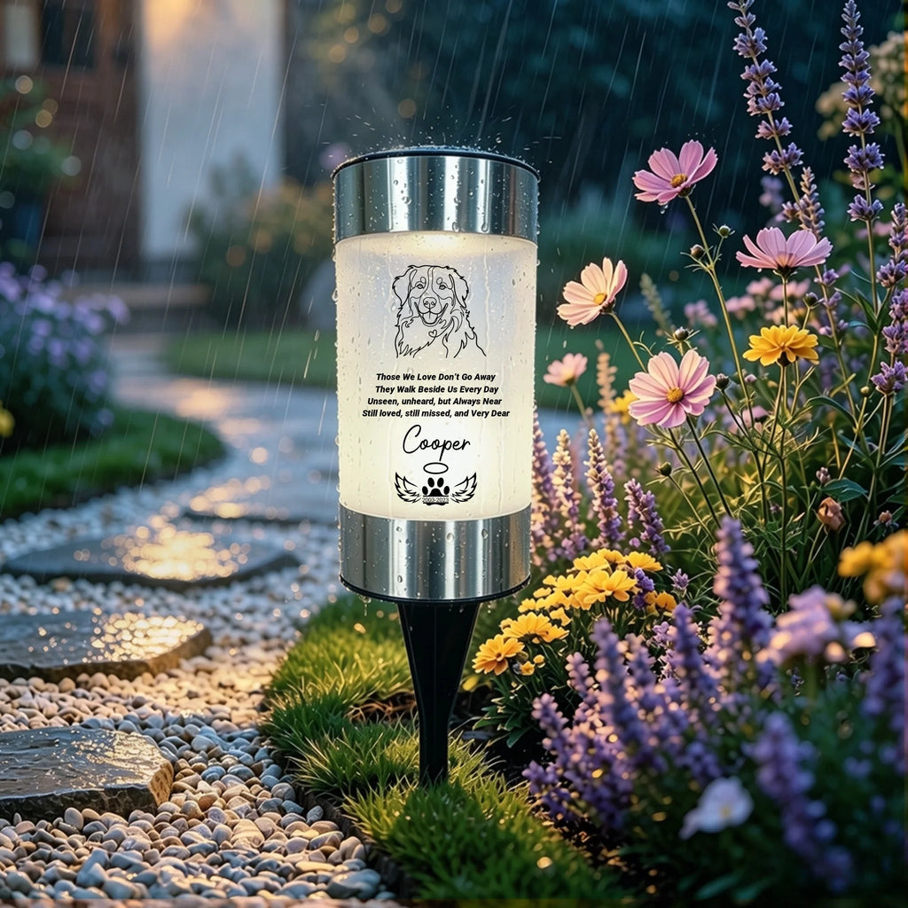 A cylindrical outdoor garden light with a silver metal top and bottom and a glowing white center panel, mounted on a black stake in a garden path. The panel shows a dog illustration, a quote, and the name 'Casper'.