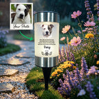 A custom pet memorial garden light shown in a rainy garden setting. The cylindrical light has a silver metal top and bottom, a glowing white center, and a black stake.