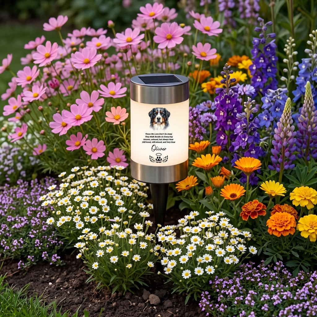 A solar-powered garden light shaped like a candle stands in a colorful flower bed. The candle-style light has a black top panel, a silver metal base, and a warm white glowing front with a dog portrait and printed text.