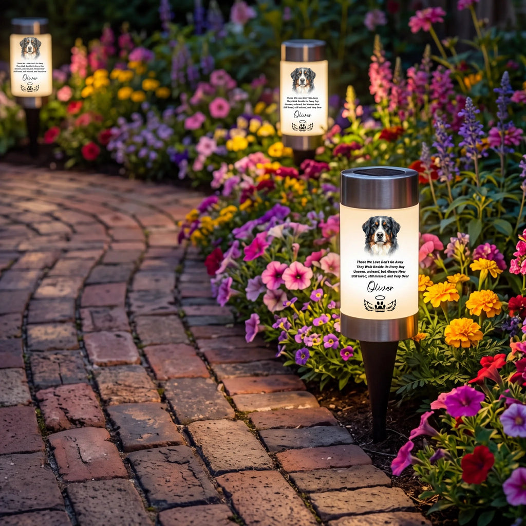 Three custom solar garden lights with black metal stakes are shown along a brick pathway next to a vibrant flower bed. The lights feature a warm white glowing panel with a dog watercolor portrait and memorial text.