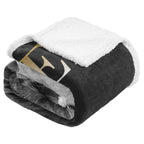 A folded fleece blanket featuring a black plush exterior with a large gold and white stylized logo, a white sherpa lining on the top, and a gray sherpa lining visible on the underside, set against a plain white background.