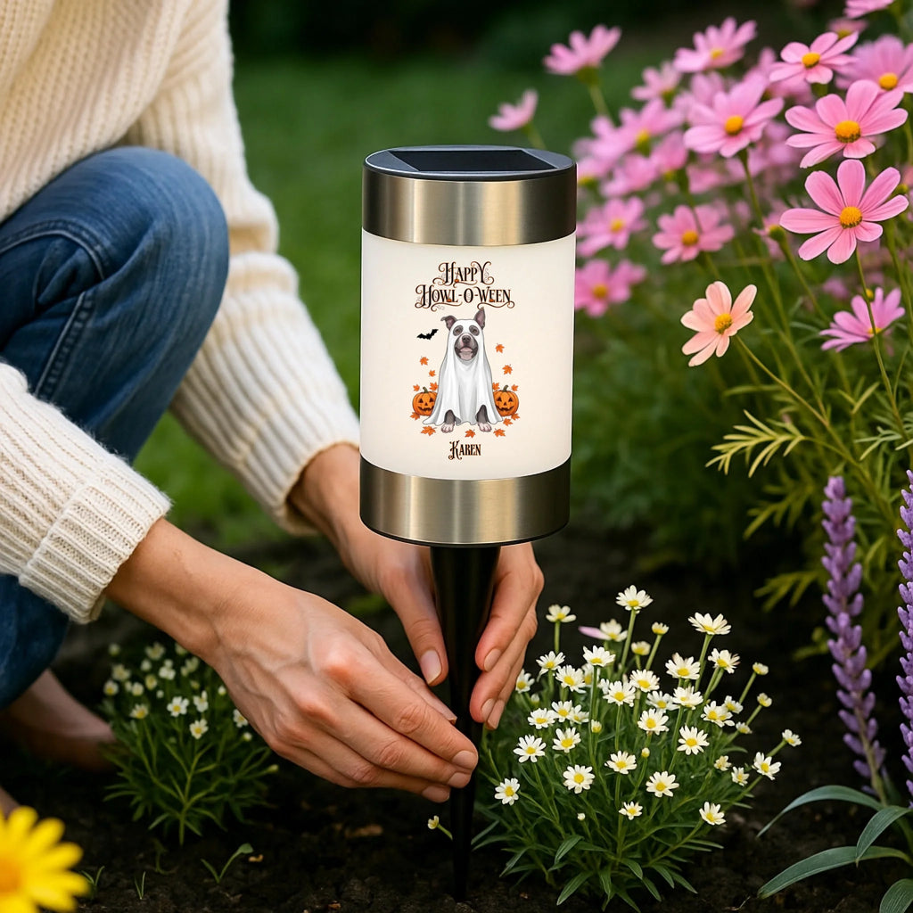 A person kneeling in a garden beside a solar-powered dog garden light. The cylindrical light has a white front panel with a printed dog illustration and the words 'Happy Halloween' and '2024', with silver metal top and bottom bands and a black stake.