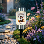 A cylindrical outdoor garden light with a black stake stands on grass beside a stone path and flowering plants in the rain. The light is illuminated and printed with a dog illustration, pumpkins, bats, and the text “Happy Home Owner” and “Jenyn.