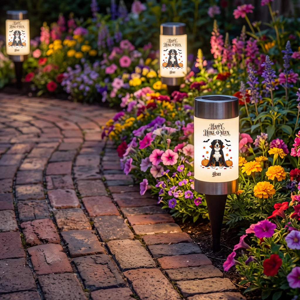 Three decorative outdoor solar garden lights with a 'Happy Halloween' dog design are shown along a brick pathway next to a vibrant flower bed.