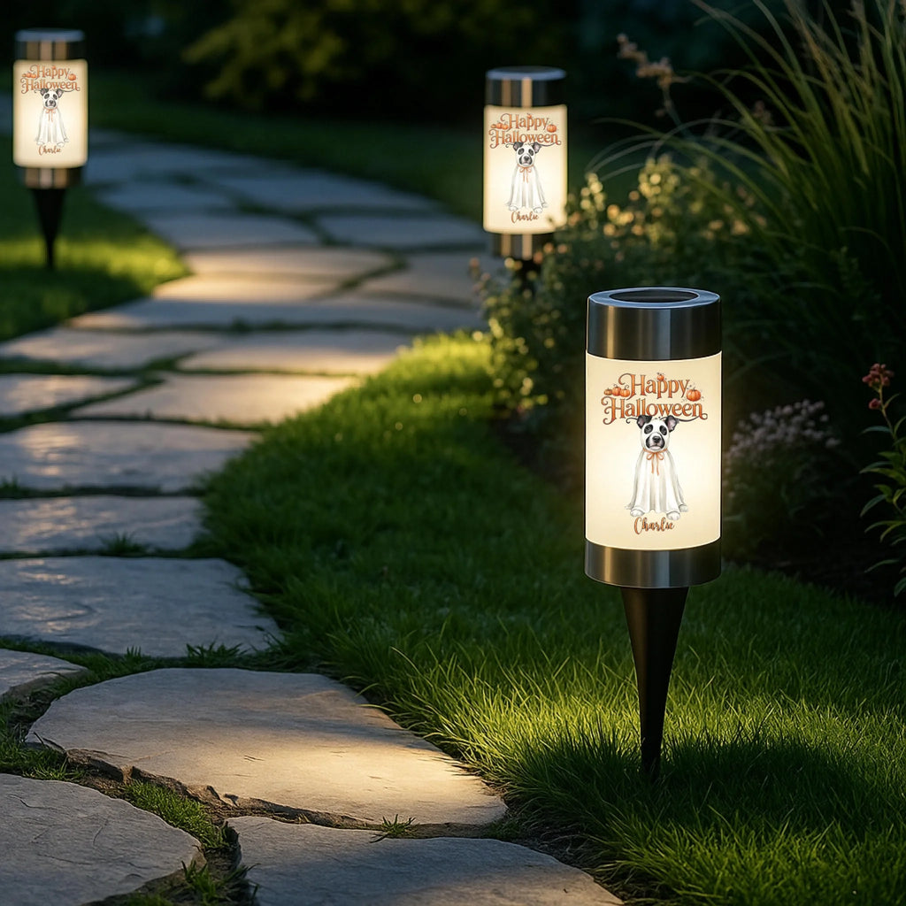 Three outdoor solar garden lights with cylindrical white shades are placed along a stone path in a landscaped garden. The lights are turned on, casting a warm glow, and feature a printed design of a black-and-white dog in a wedding gown with the text 'Happy Halloween' and 'Love, Clara.