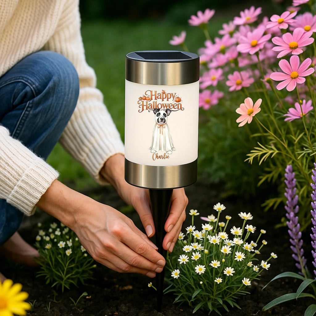 A person kneeling in a garden beside a solar-powered stake light with a frosted white body, black top and bottom bands, and a printed Halloween illustration that reads 'Happy Halloween Uncle'. The scene is outdoors with flowers and greenery in the background.