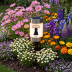 A solar-powered garden stake light with a glowing 'Happy Halloween' witch graphic, set in a colorful flower bed surrounded by pink, purple, white, and orange flowers.