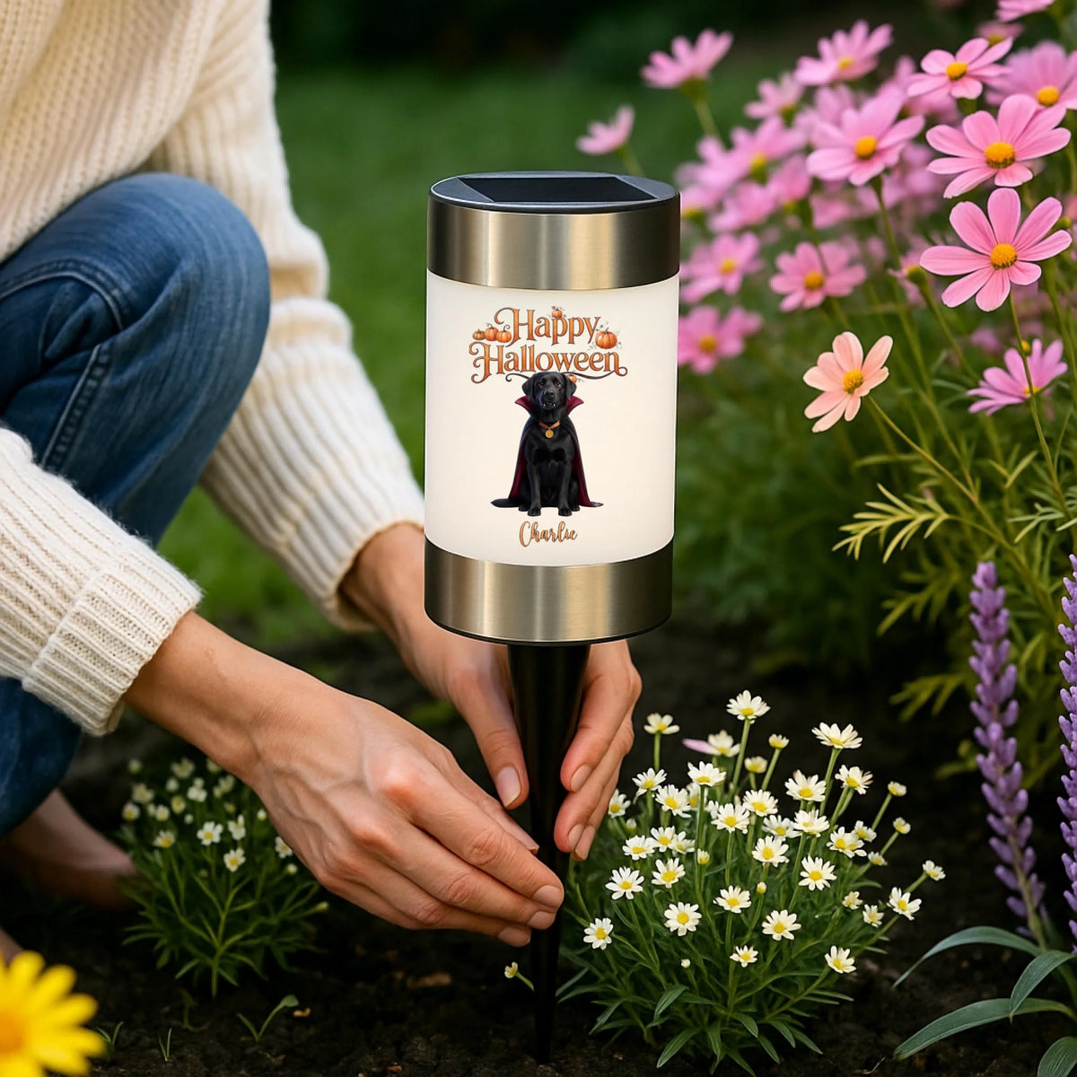 A person kneeling in a garden beside a solar-powered Halloween garden stake light. The cylindrical light has a white label with 'Happy Halloween' and a black cat illustration, plus the name 'Charlie' at the bottom.