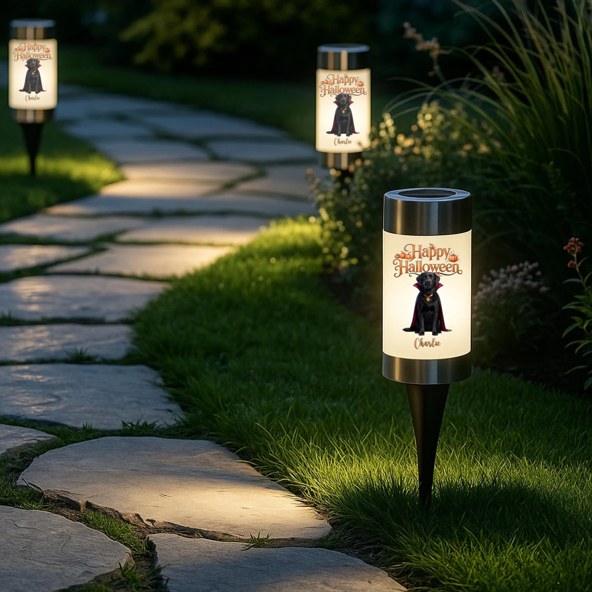 Three outdoor solar pathway lights shaped like vampire skulls are mounted on grass beside a stone garden path at night. The lights glow with a white background and feature a black skull illustration with the words 'Happy Halloween' and the name 'Chucky' printed below.
