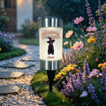 A cylindrical solar garden light with a warm white glow, featuring a custom 'Happy Halloween' design with a black cat and the name 'Charlie', set in a garden path during a rainy evening.