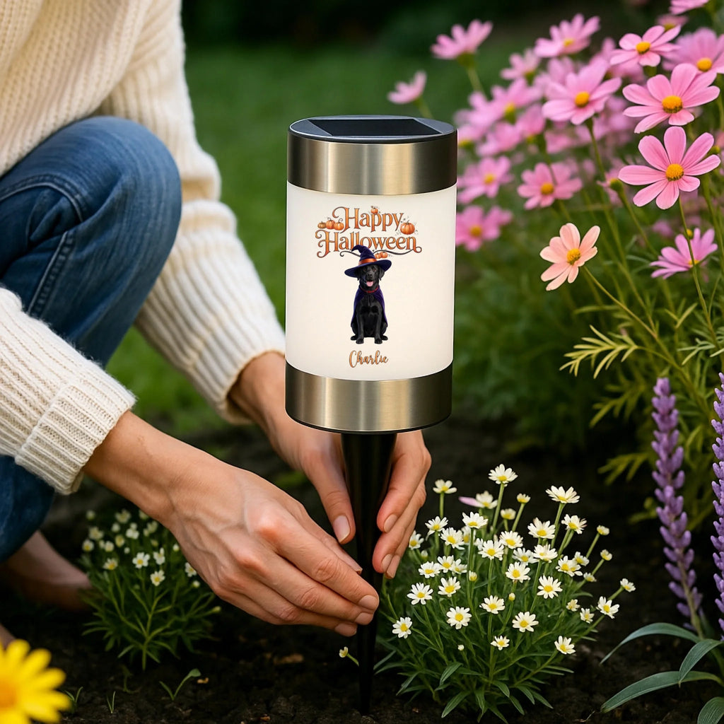 A person kneeling in a garden beside a cylindrical solar-powered garden light stake. The light has a white body with a printed 'Happy Halloween' design featuring a witch and the name 'Charlie', with a black stake and silver top and bottom bands.