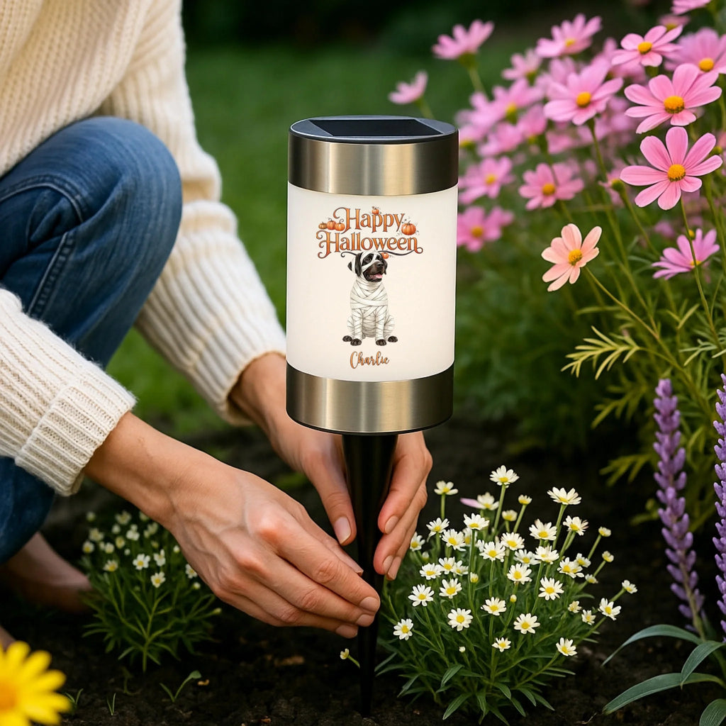 A person kneeling in a garden beside a solar-powered dog garden light. The cylindrical light has a white front panel with a printed dog illustration, a personalized dog photo transformed into a mummy, and the words 'Happy Halloween'  with silver metal top and bottom bands and a black stake.