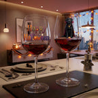 Two personalized wine glasses filled with red wine on a dining table, decorated with 'Happy Halloween' text and a small black dog wearing a vampire costume. The background shows a cozy, warmly lit dining room with Halloween decorations.