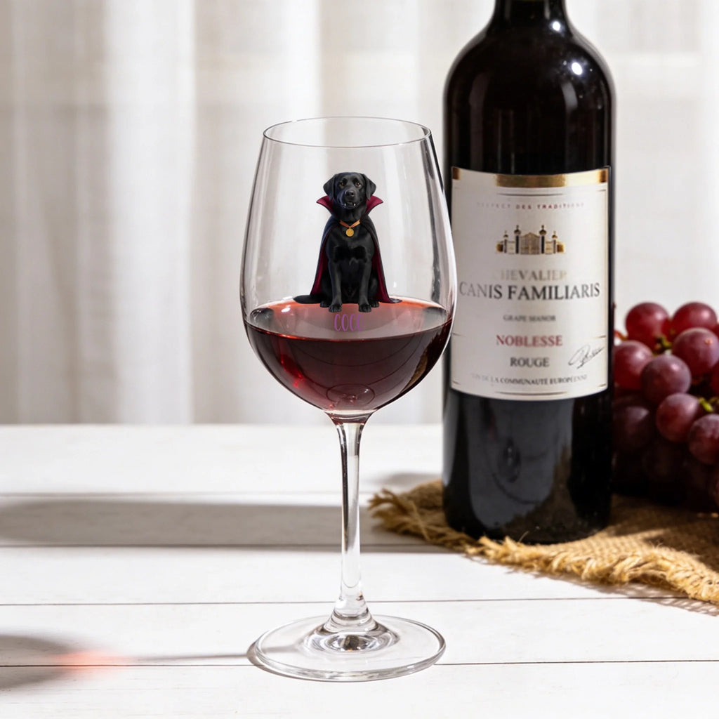 A clear wine glass partially filled with red wine, featuring a printed black dog wearing a red cape and gold collar, set on a white tabletop. A dark wine bottle and a bunch of red grapes sit in the softly lit background.