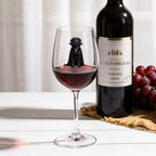 A clear wine glass partially filled with red wine, featuring a printed black dog wearing a red cape and gold collar, set on a white tabletop. A dark wine bottle and a bunch of red grapes sit in the softly lit background.