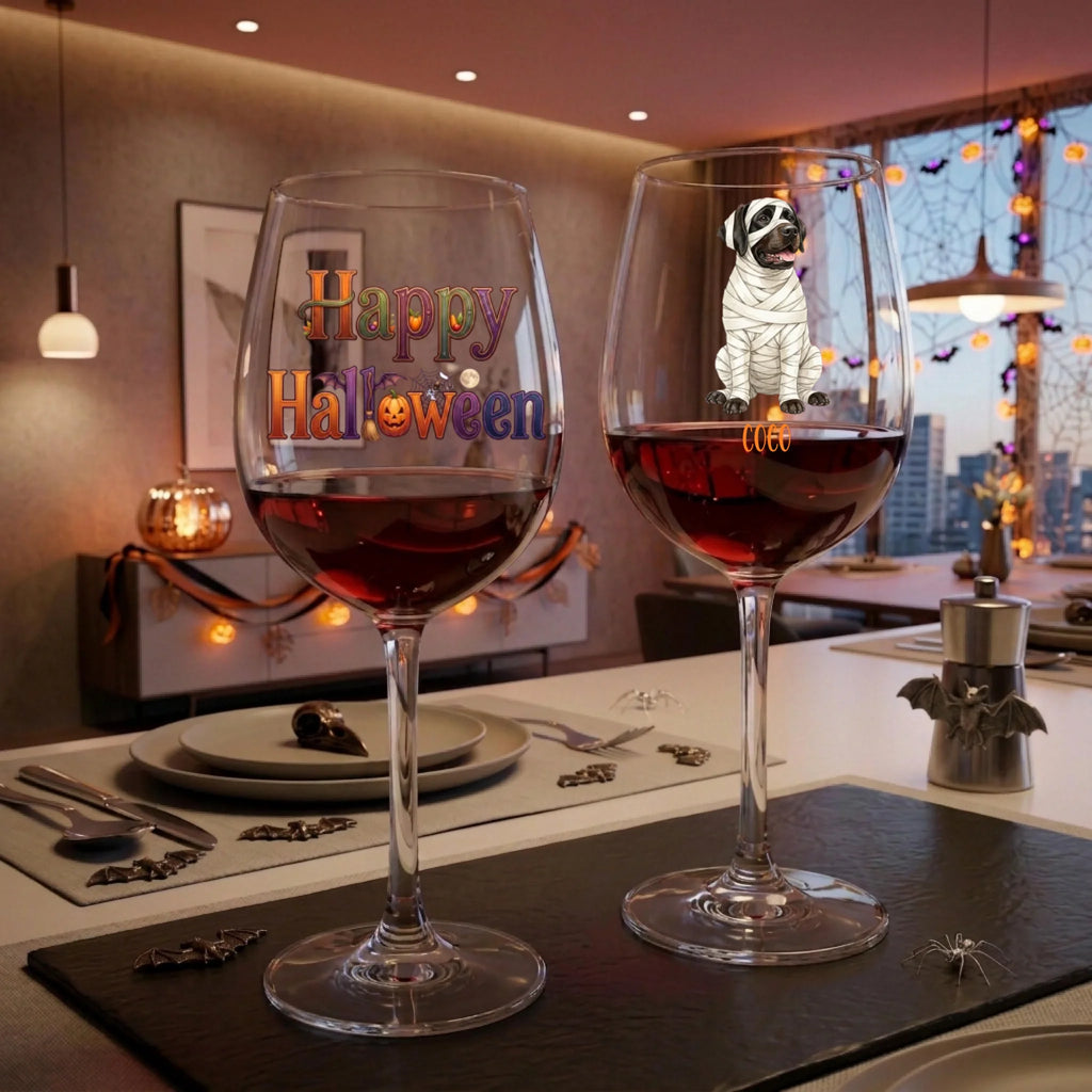 Two clear wine glasses filled with red wine on a dining table, decorated with Halloween-themed graphics. The left glass has 'Happy Halloween' in colorful letters, and the right glass features a cartoon mummy dog.