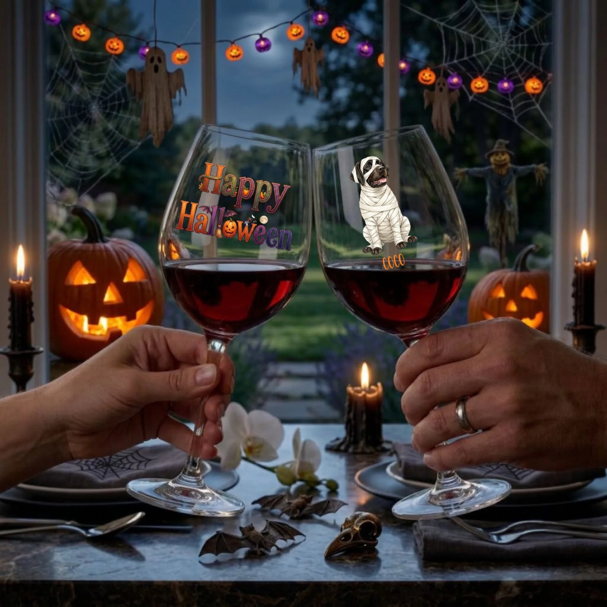 Two hands holding wine glasses filled with red wine at a Halloween-themed dinner table. The left glass is printed with 'Happy Halloween' in orange and purple lettering, and the right glass features a cartoon mummy dog.
