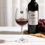 A clear wine glass with a small dog figurine inside, partially filled with red wine, set on a white tabletop next to a dark wine bottle and a bunch of red grapes.