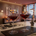 Two stemmed wine glasses filled with red wine are displayed on a dining table in a decorated indoor setting. The left glass has colorful 'Happy Halloween' lettering, and the right glass features a mummy dog illustration with the name 'NICOLY' printed below.