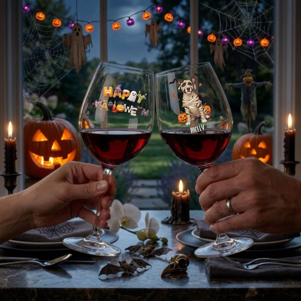 Two hands hold up clear wine glasses filled with red wine at a Halloween dinner table. The left glass has colorful 'Happy Halloween' lettering, and the right glass features a cartoon dog wearing a tattered superhero cape with 'Molly' written below.