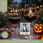 Two personalized stemmed wine glasses filled with red wine, decorated with Halloween-themed graphics including 'Happy Halloween' and a dog mummy design. The glasses are shown on a wooden table with a pumpkin, berries, and a photo insert in a warm, festive setting.