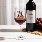 A clear wine glass with a black line drawing of a French Bulldog and the name 'Frankie" printed on it, filled with red wine, set on a white tabletop next to a dark wine bottle and a bunch of red grapes.