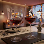 Two Halloween-themed wine glasses filled with red wine, set on a dining table decorated for the holiday. The left glass features 'HAPPY HALLOWEEN' with ghosts and pumpkins,