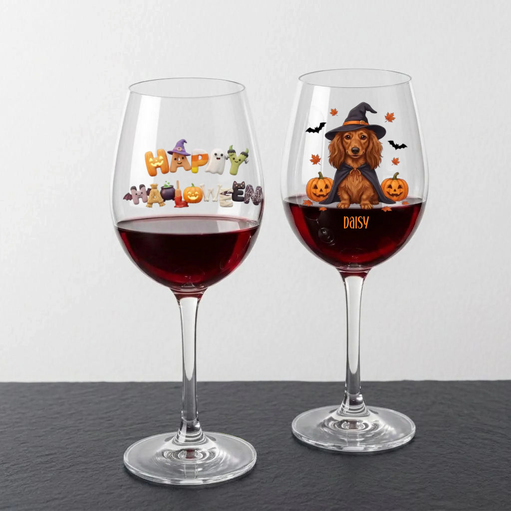 Two clear wine glasses filled with red wine, displayed on a dark slate surface against a light gray wall. The left glass has a colorful 'Happy Halloween' design, and the right glass is personalized with the name 'DAISY' below an illustration of a dachshund wearing a witch hat with pumpkins and bats.