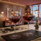 Two Halloween-themed wine glasses filled with red wine on a dining table. One glass has 'Happy Halloween' with spooky cutout letters, and the other has a dachshund wearing a witch hat surrounded by pumpkins with the name 'Daisy' written below.