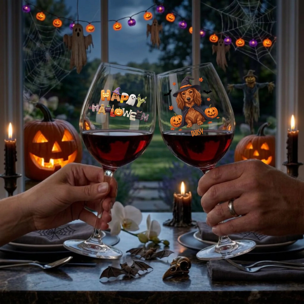 Two hands hold up clear wine glasses filled with red wine at a Halloween table setting. The left glass is printed with 'Happy Halloween' in orange and yellow letters, and the right glass features a cartoon dog wearing a witch hat with orange pumpkins.