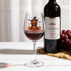 A clear wine glass filled with red wine, decorated with a cartoon dachshund wearing a witch hat and surrounded by pumpkins and bats, set on a white table next to a wine bottle and a bunch of red grapes.
