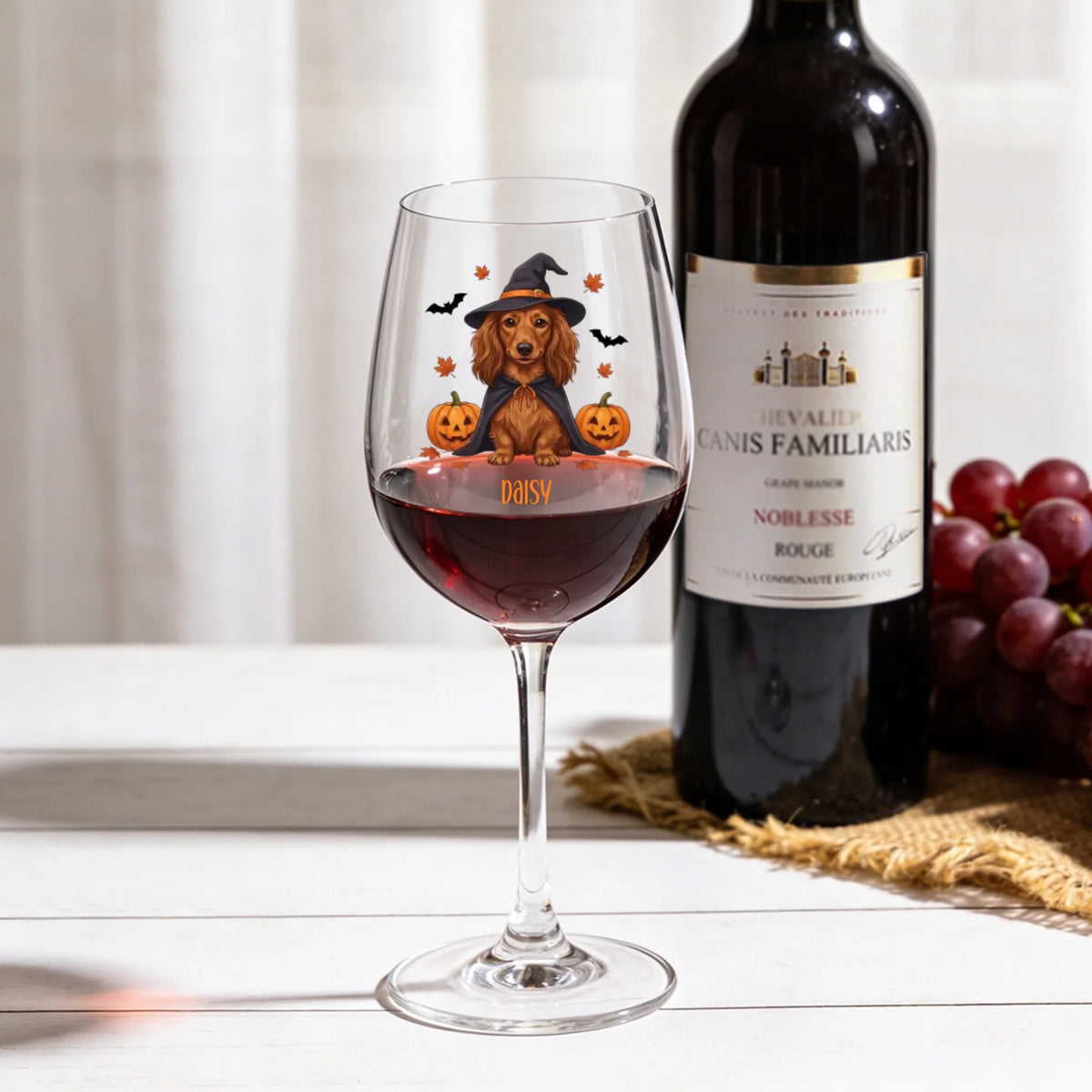 A clear wine glass filled with red wine, decorated with a cartoon dachshund wearing a witch hat and surrounded by pumpkins and bats, set on a white table next to a wine bottle and a bunch of red grapes.