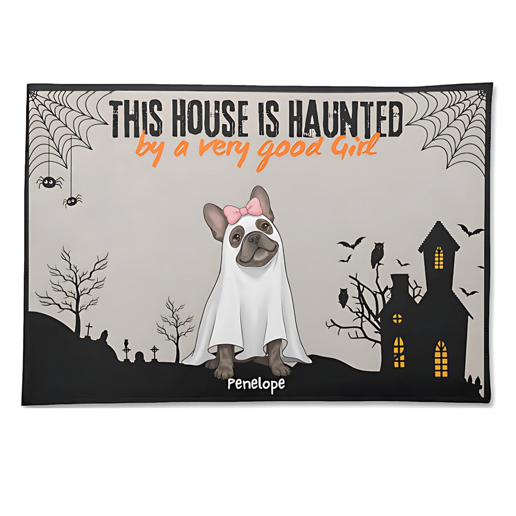 A rectangular Halloween-themed mat or rug with a printed design featuring a French bulldog dressed as a ghost, the text 'THIS HOUSE IS HAUNTED by a very good Girl', a haunted house, spiderwebs, bats, and the name 'Penelope' at the bottom. The mat is shown against a plain white background.