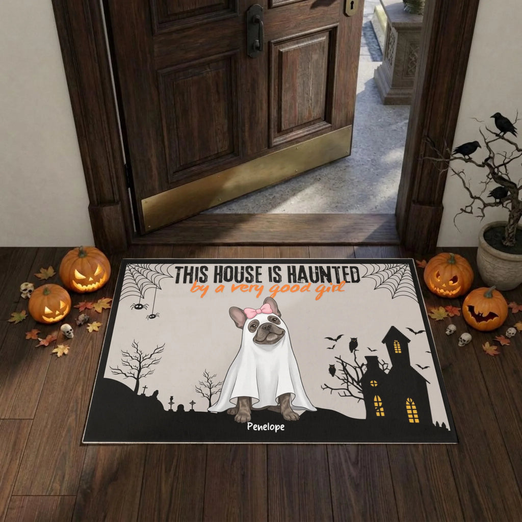 A Halloween-themed doormat displayed in front of a wooden door on a wooden porch. The mat features a cartoon French Bulldog dressed as a ghost with the text 'THIS HOUSE IS HUAUNTED by a very good gut' and the name 'P envelope'.