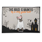 A rectangular Halloween-themed mat or rug with a printed design featuring a French bulldog dressed as a ghost, the text 'THIS HOUSE IS HAUNTED by a very good Girl', a haunted house, spiderwebs, bats, and the name 'Penelope' at the bottom. The mat is shown against a plain white background.