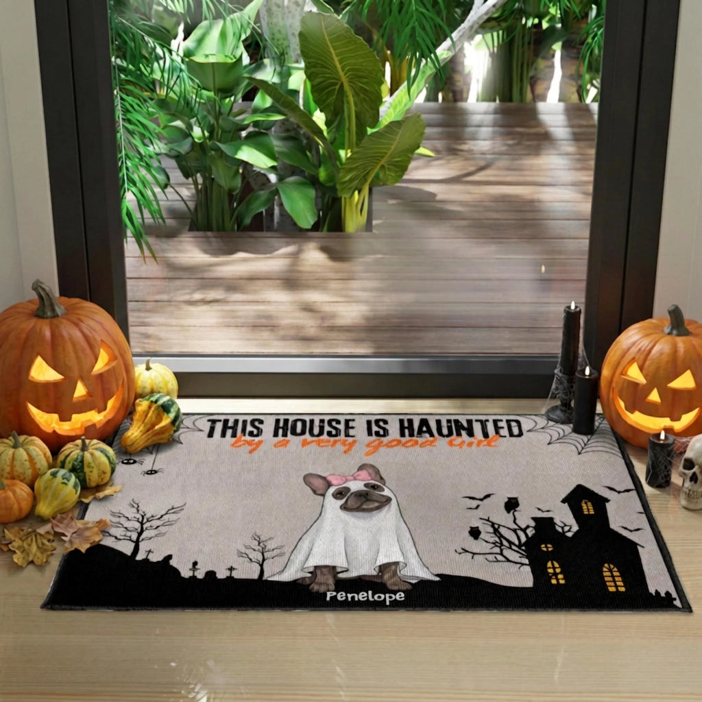 A Halloween-themed doormat featuring a French bulldog in a ghost costume, set in front of a glass door with pumpkins and fall decorations around it.