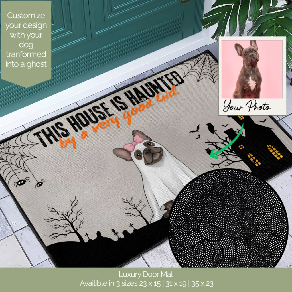 A Halloween-themed doormat shown on a tiled entryway floor in front of a teal door. The mat features the text 'THIS HOUSE IS HAUNTED by a very good cat' with a ghost illustration, spiderwebs, bats, and a haunted house.