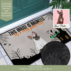 A Halloween-themed doormat shown on a tiled entryway floor in front of a teal door. The mat features the text 'THIS HOUSE IS HAUNTED by a very good cat' with a ghost illustration, spiderwebs, bats, and a haunted house.