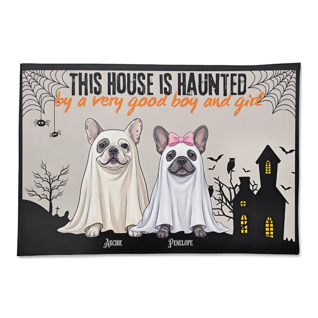 A rectangular Halloween-themed doormat with a black border, featuring two French bulldogs in white ghost costumes, a haunted house, bats, spiders, and the text 'THIS HOUSE IS HAUNTED by a very good boy and girl' above the dogs.