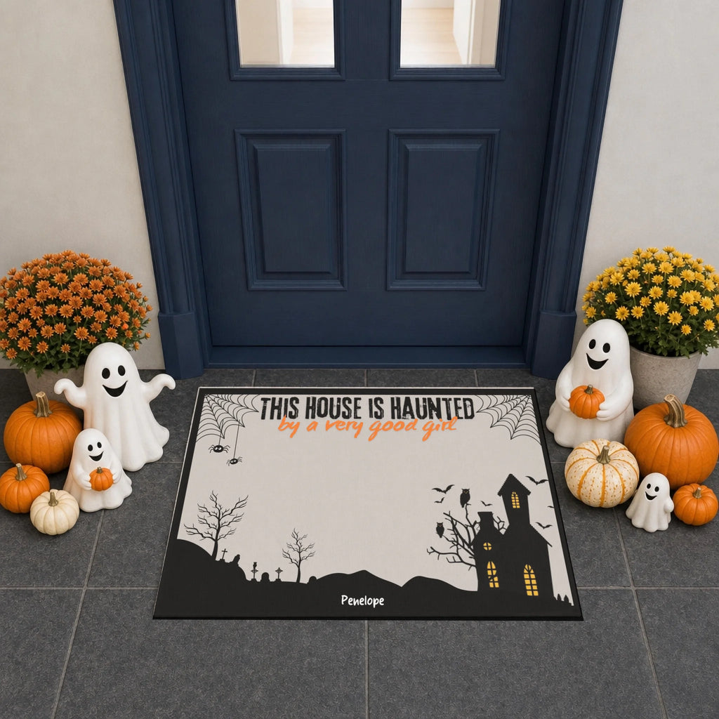 A Halloween-themed doormat featuring a cartoon French Bulldog in a ghost costume with the text 'THIS HOUSE IS HUAUNTED by a very good gut'. The mat is displayed on a gray tiled entryway floor in front of a dark blue door, surrounded by decorative pumpkins, ghost figurines, and yellow flowers.