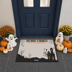A Halloween-themed doormat featuring a cartoon French Bulldog in a ghost costume with the text 'THIS HOUSE IS HUAUNTED by a very good gut'. The mat is displayed on a gray tiled entryway floor in front of a dark blue door, surrounded by decorative pumpkins, ghost figurines, and yellow flowers.