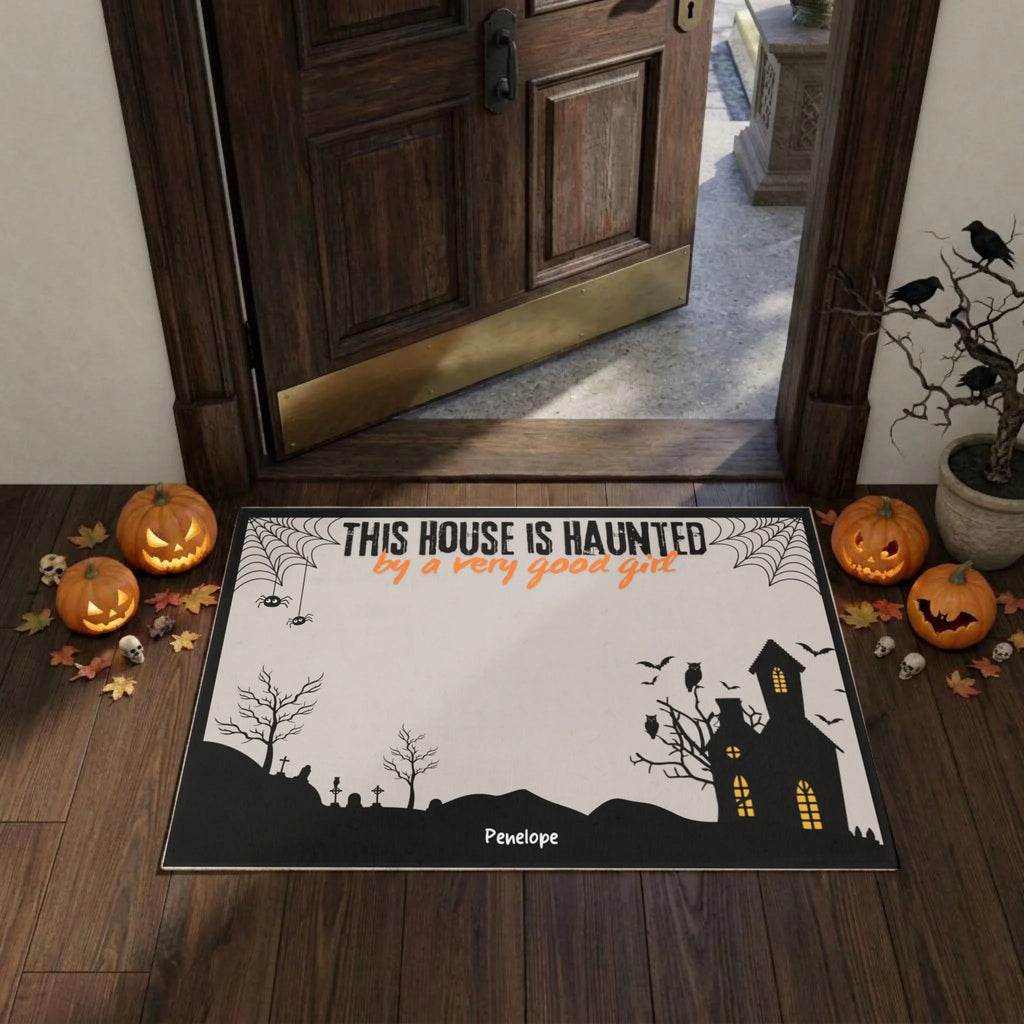 A Halloween-themed doormat displayed on a dark wood entryway floor in front of an open wooden door. The mat features a black silhouette of a haunted house, bats, spiderwebs, and tombstones on an off-white background, with the text 'THIS HOUSE IS HAUNTED by a very good grot' in black and orange.