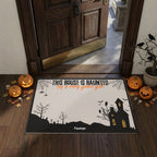A Halloween-themed doormat displayed on a dark wood entryway floor in front of an open wooden door. The mat features a black silhouette of a haunted house, bats, spiderwebs, and tombstones on an off-white background, with the text 'THIS HOUSE IS HAUNTED by a very good grot' in black and orange.