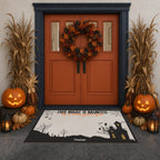 A Halloween-themed doormat featuring a cartoon French Bulldog in a ghost costume, placed in front of a dark blue double door decorated with a wreath, jack-o'-lanterns, and cornstalks.