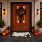 A Halloween-themed doormat with a spooky black and white haunted house design and the text 'THIS HOUSE IS HAUNTED' is displayed in front of a double orange-brown door decorated with a wreath, pumpkins, and lanterns.