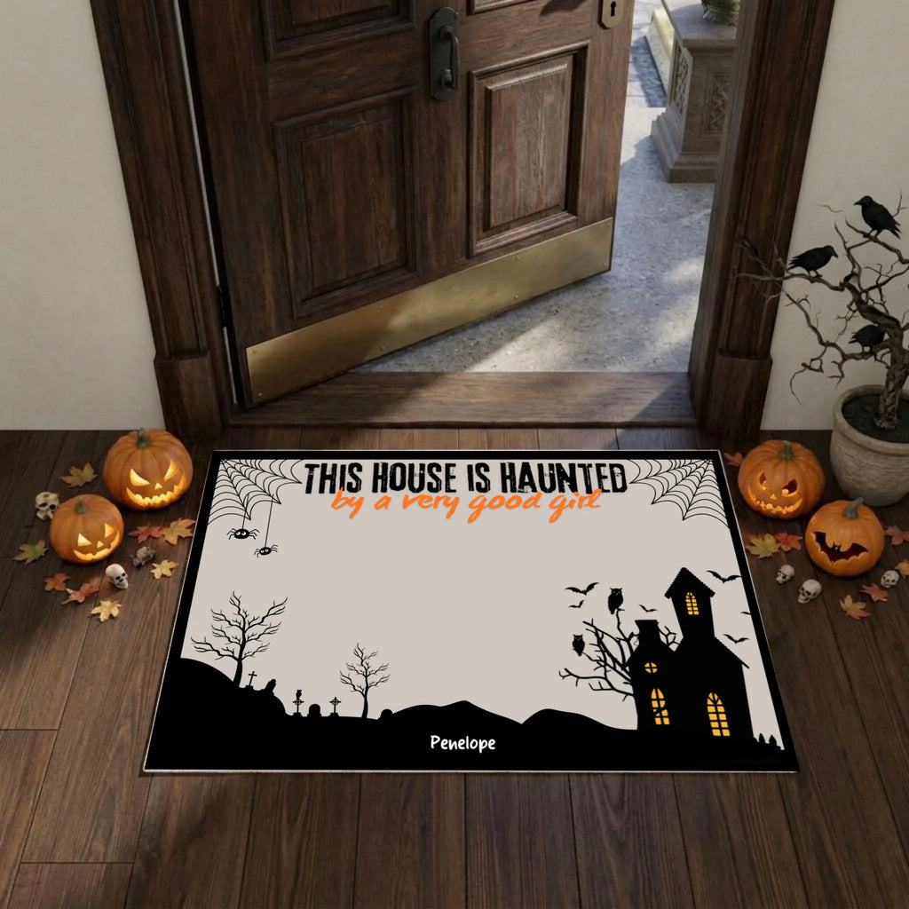 A Halloween-themed doormat displayed on a wooden entryway floor in front of an open dark wood door. The mat features a black silhouette landscape with a haunted house, bats, spiderwebs, and the text 'THIS HOUSE IS HAUNTED by a very good gait' on an off-white background.