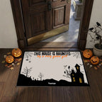 A Halloween-themed doormat displayed on a wooden entryway floor in front of an open dark wood door. The mat features a black silhouette landscape with a haunted house, bats, spiderwebs, and the text 'THIS HOUSE IS HAUNTED by a very good gait' on an off-white background.